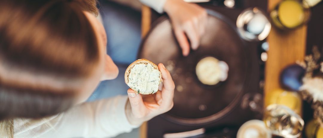 Eine Person hält ein Sandwich mit grüner Aufstrich, schaut auf einen Tisch mit einer Pfanne und anderen Lebensmitteln.