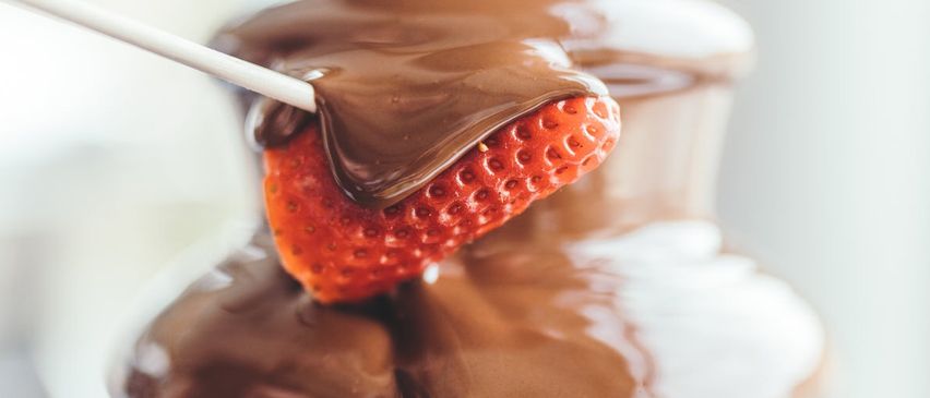 Eine Schokoladenfontäne mit einer in Schokolade getauchten Erdbeere, die an einem Stiel gehalten wird. Die Fontäne hat einen glatten Schokoladenfluss.