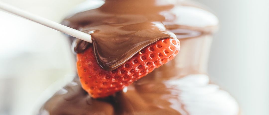 Eine Erdbeere wird in Schokoladensauce aus einer Schokoladenfontäne getaucht. Die Erdbeere ist auf einem Stiel und hat einen glänzenden Finish.