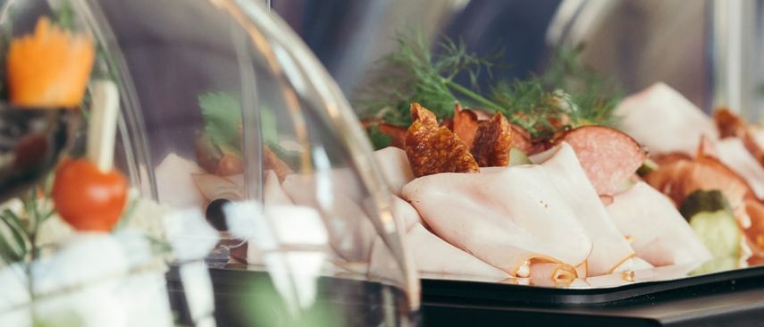 Ein Tisch mit verschiedenen Speisen unter Glaskuppeln. Es gibt Fleischscheiben, Gemüse und Früchte. Links ist eine Salatschüssel zu sehen.