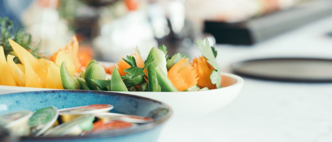 A blue bowl filled with a colorful salad sits on a table, with blurry food in the background.