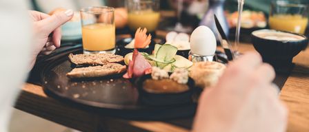 Ein Frühstück auf einem Holztisch mit einem Glas Orangensaft, Brot, Eiern und Gemüse. Eine Hand hält Messer und Gabel.