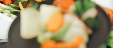A plate filled with chopped vegetables, including carrots and cucumbers, is being lifted by a hand. A tong is on a plate with a piece of meat.