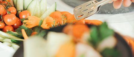 A person holds a plate of sliced vegetables with a fork in hand, with a bowl of tomatoes and cucumbers in the background.
