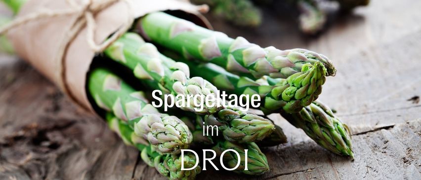Ein Bündel frischer grüner Spargelstangen, eingewickelt in braunes Papier, mit dem Text 'Spargeltage im DR0I' darüber. Der Spargel hat ein leicht glänzendes, wachsartiges Aussehen.