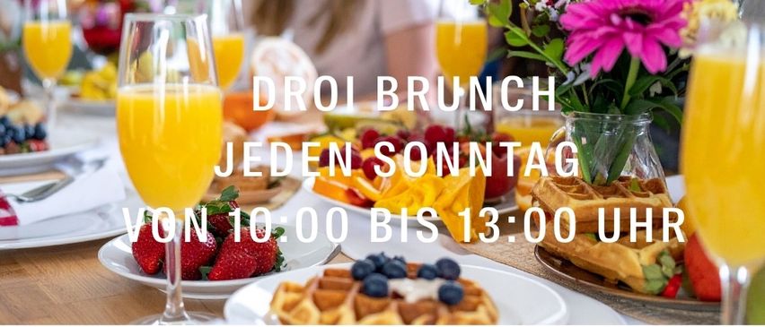 Ein Brunchtisch ist mit verschiedenen Speisen und Getränken gedeckt. Gläser mit Orangensaft stehen auf dem Tisch. Der Text 'DROI BRUNCH JEDEN SONNTAG VON 10:00 BIS 13:00 UHR' ist angezeigt.