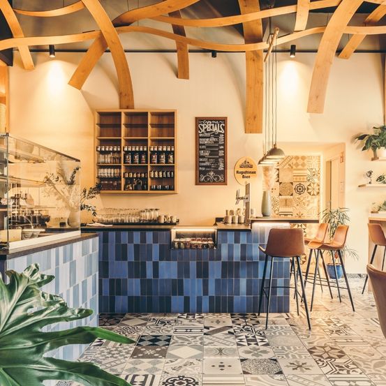 Ein gemütliches Café-Interieur mit Holzbalken, blau gefliester Theke, Regalen mit Flaschen, einem Kreidetafel mit Spezialitäten und einer gemusterten Wand. Es gibt auch Pflanzen und Hängelampen.