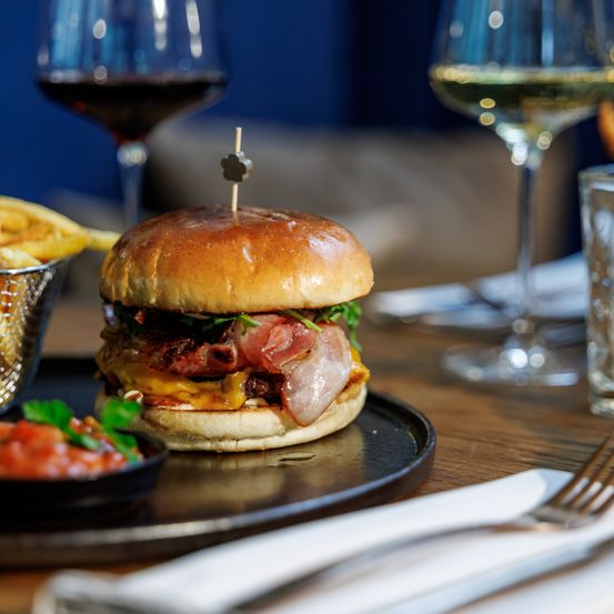 Ein Cheeseburger mit Speck und geschmolzenem Käse auf einem Brötchen, begleitet von einem Glas Wein und Pommes auf einem Holztisch.