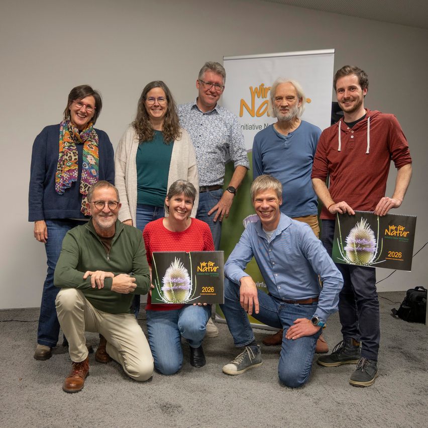 Eine Gruppe von Menschen posiert für ein Foto in einem Raum. Sie halten ein Banner mit den Worten 'Wir für Natur'. Sie alle lächeln und einige von ihnen halten ein Poster mit einem Bild einer Blume.