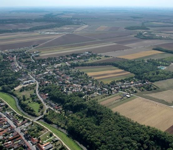 Luftaufnahme eines ländlichen Gebiets mit einer kleinen Stadt, die von vielen Gebäuden umgeben ist. Sie wird von weitläufigen Feldern und Bäumen gesäumt. Die Landschaft wird von landwirtschaftlichen Flächen dominiert, mit Flächen unterschiedlicher Nutzpflanzen und einem Fluss, der durch die Stadt fließt.