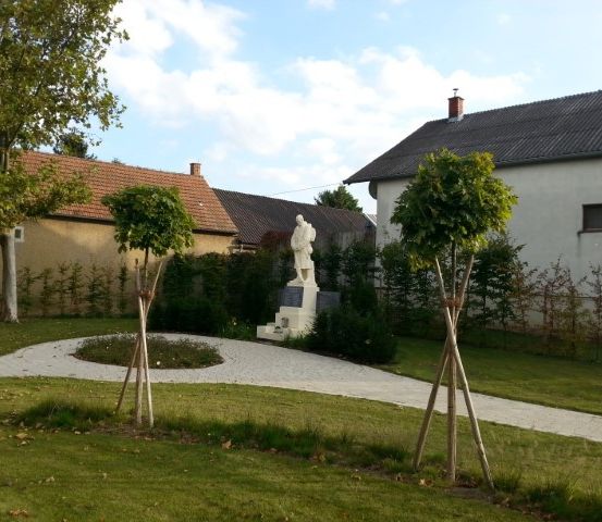 Ein Garten mit einer Statue, umgeben von grünem Rasen, Bäumen und Büschen. Hinter der Statue stehen zwei Häuser mit Schornsteinen.