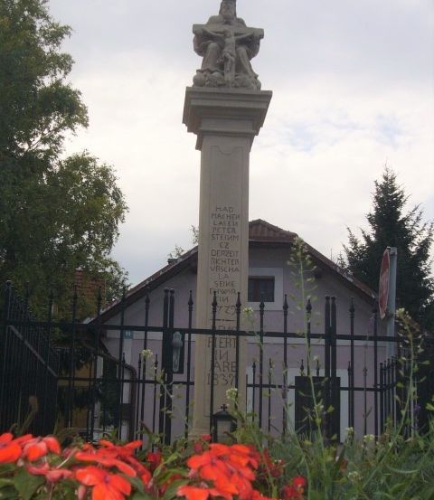 Eine Statue von Jesus am Kreuz steht auf einem Steinpfeiler vor einem Haus mit roten Blumen vor einem Zaun.