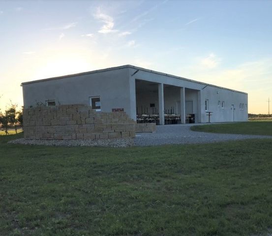 Ein modernes Gebäude mit einer Ziegelmauer steht in einem grünen Feld. Mehrere Fenster und eine Tür sind sichtbar. In der Ferne sind Windturbinen und Bäume zu sehen.