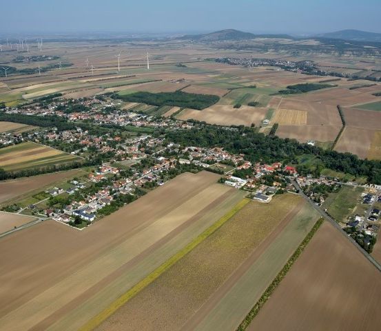 Luftaufnahme einer ländlichen Gegend mit Feldern, Häusern und Bäumen. In der Ferne sind Berge zu sehen, und Windturbinen sind sichtbar.