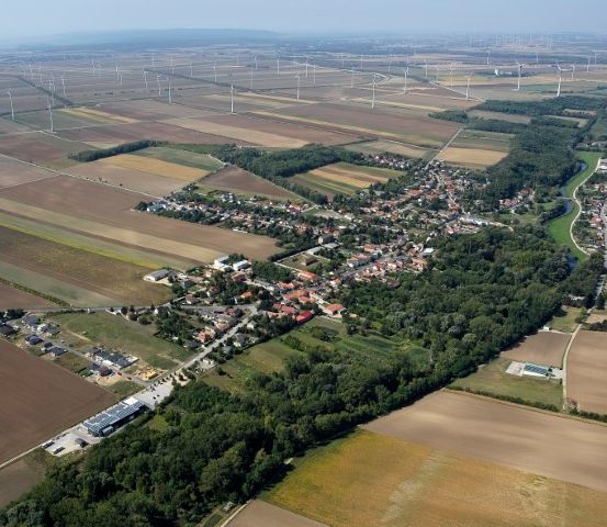 Eine Luftaufnahme eines ländlichen Gebiets mit zahlreichen Feldern, einer Stadt in der Mitte und mehreren Windturbinen in der Ferne.
