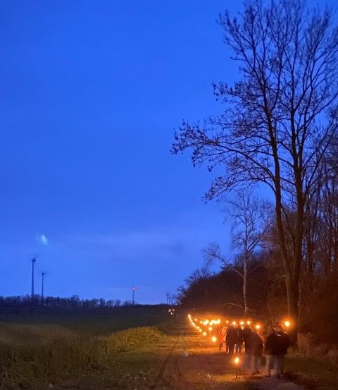 Eine Gruppe von Menschen geht bei Dämmerung einen mit brennenden Kerzen gesäumten Pfad in einem ländlichen Gebiet entlang.