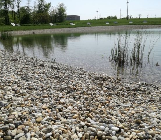 Ein See mit Kiesstrand, umgeben von Grün und einem Gebäude in der Ferne.