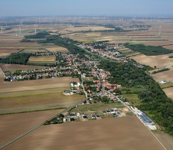 Luftaufnahme einer kleinen Stadt mit mehreren Häusern, einer kurvenreichen Straße und weitläufigen Feldern. Zahlreiche Windräder säumen den Horizont.