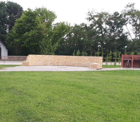 Eine Steinbank in einem Rasenfeld mit einer kleinen Kapelle und Bäumen im Hintergrund. Ein Backsteinkamin befindet sich rechts von der Bank.