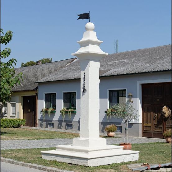 Ein weißer Denkmal mit schwarzer Fahne steht vor einem Gebäude mit grauem Dach und grünen Fenstern. Zwei Topfpflanzen stehen auf beiden Seiten des Denkmals.