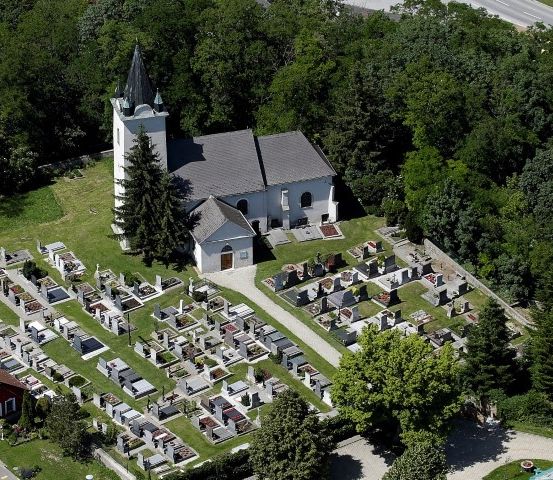 Ein Luftbild einer kleinen Kirche, umgeben von einem Friedhof mit vielen Grabsteinen, in einem üppig grünen Bereich gelegen.