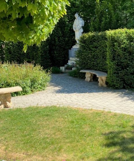 Ein ruhiger Garten mit einer Statue in der Mitte, umgeben von gepflegten Hecken und Ziegelpfaden. Zwei Steinbänke sind einladend im Schatten eines Baumes platziert.