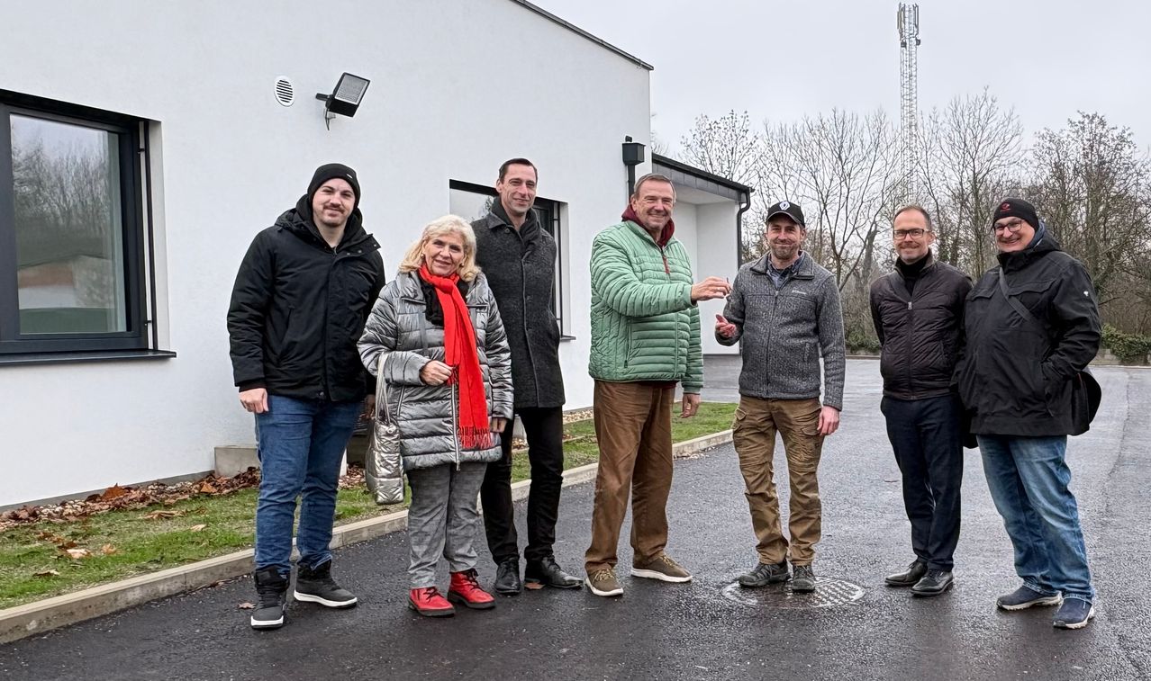 Sechs Menschen stehen vor einem Gebäude. Sie lächeln und posieren für ein Foto. Eine Frau mit rotem Schal trägt eine Tasche. Hinter ihnen steht ein Gebäude mit einer Lampe an der Wand.