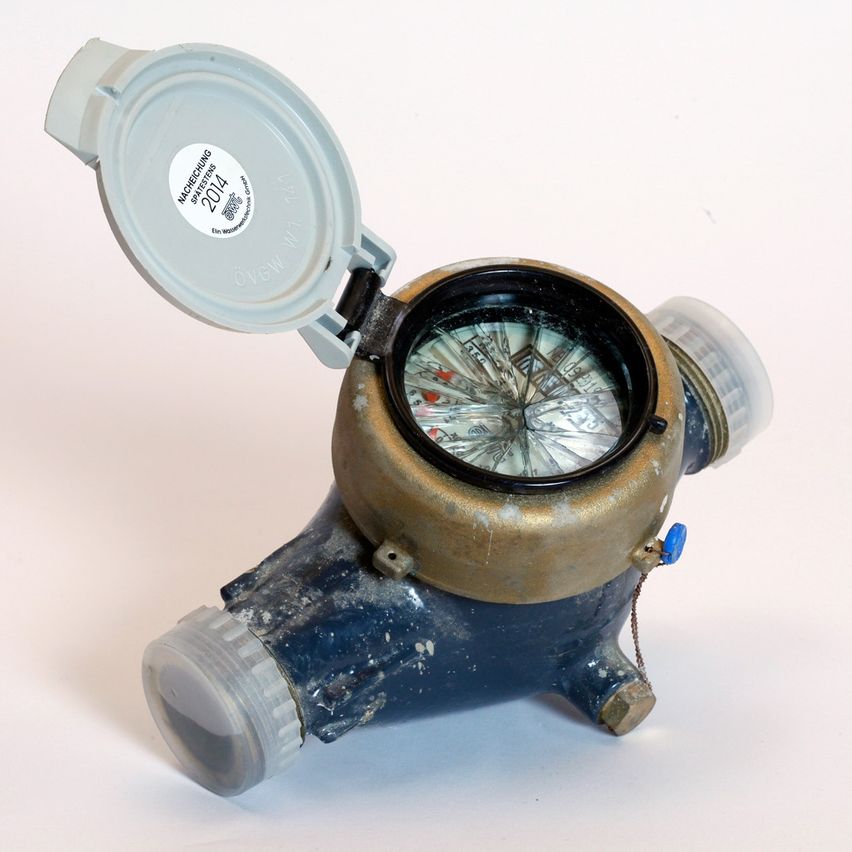 Eine Nahaufnahme eines alten Wasserzählers mit einem transparenten Deckel auf weißem Hintergrund. Der Zähler hat ein schwarzes und goldenes Farbschema, und der Deckel ist grau.