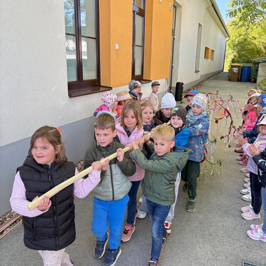 A group of children are gathered outside a building, pulling a long stick and wearing winter clothes. They are smiling and looking at the camera.