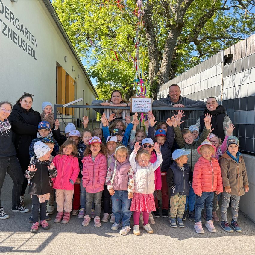 A group of children and adults pose for a photo in front of a building with a tree and a sign that reads 'Kindergarten Neusiedl'.