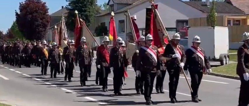 A marching band in uniforms and helmets parades down a street, carrying flags and accompanied by a white trailer.