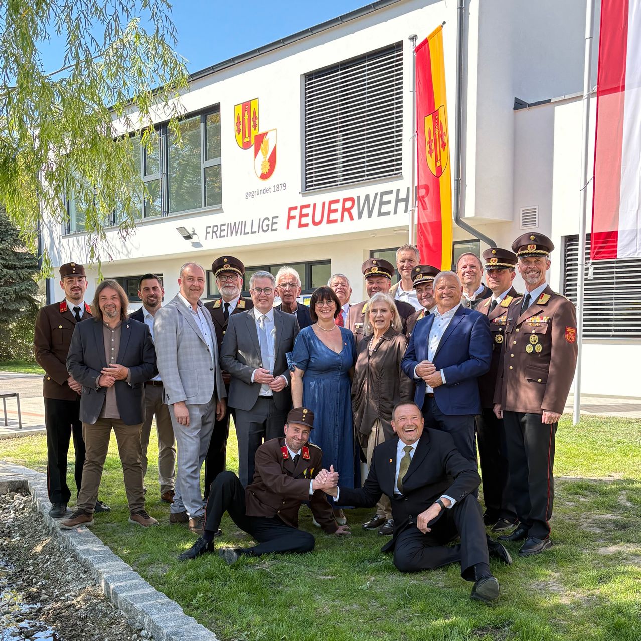 A group of people in formal attire pose for a photo in front of a building with the words Freiwillige Feuerwehr on it.