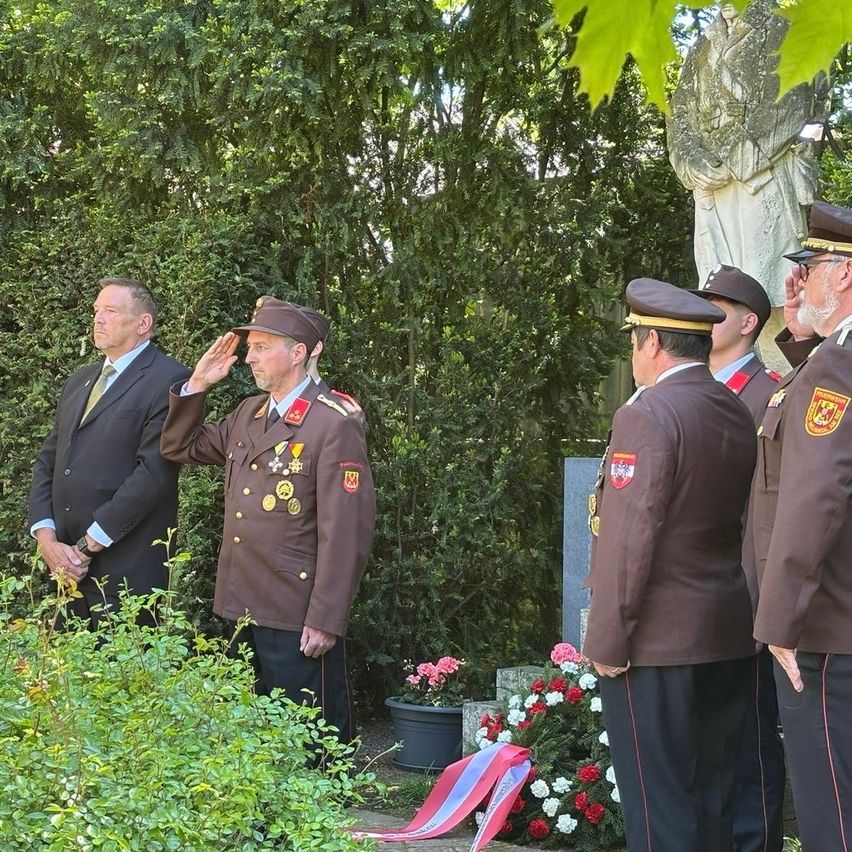 A military ceremony takes place with several officers in uniform. One officer salutes in front of a statue and a wreath.
