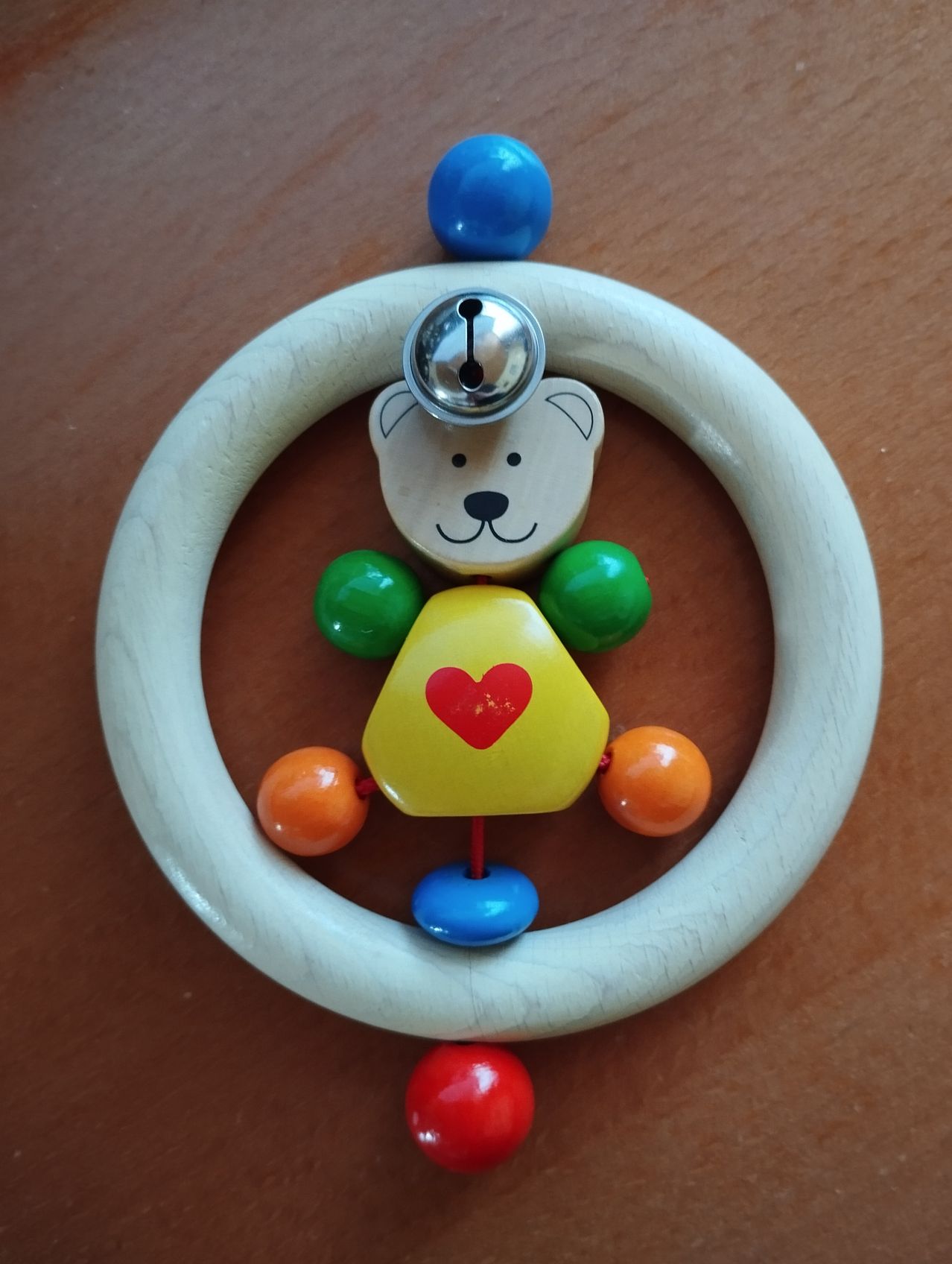 Ein holzernes Babyspielzeug zeigt einen Bären mit einem gelben Herzen auf der Brust. Das Spielzeug hat eine silberne Glocke auf der Oberseite und ist von bunten Perlen umgeben.