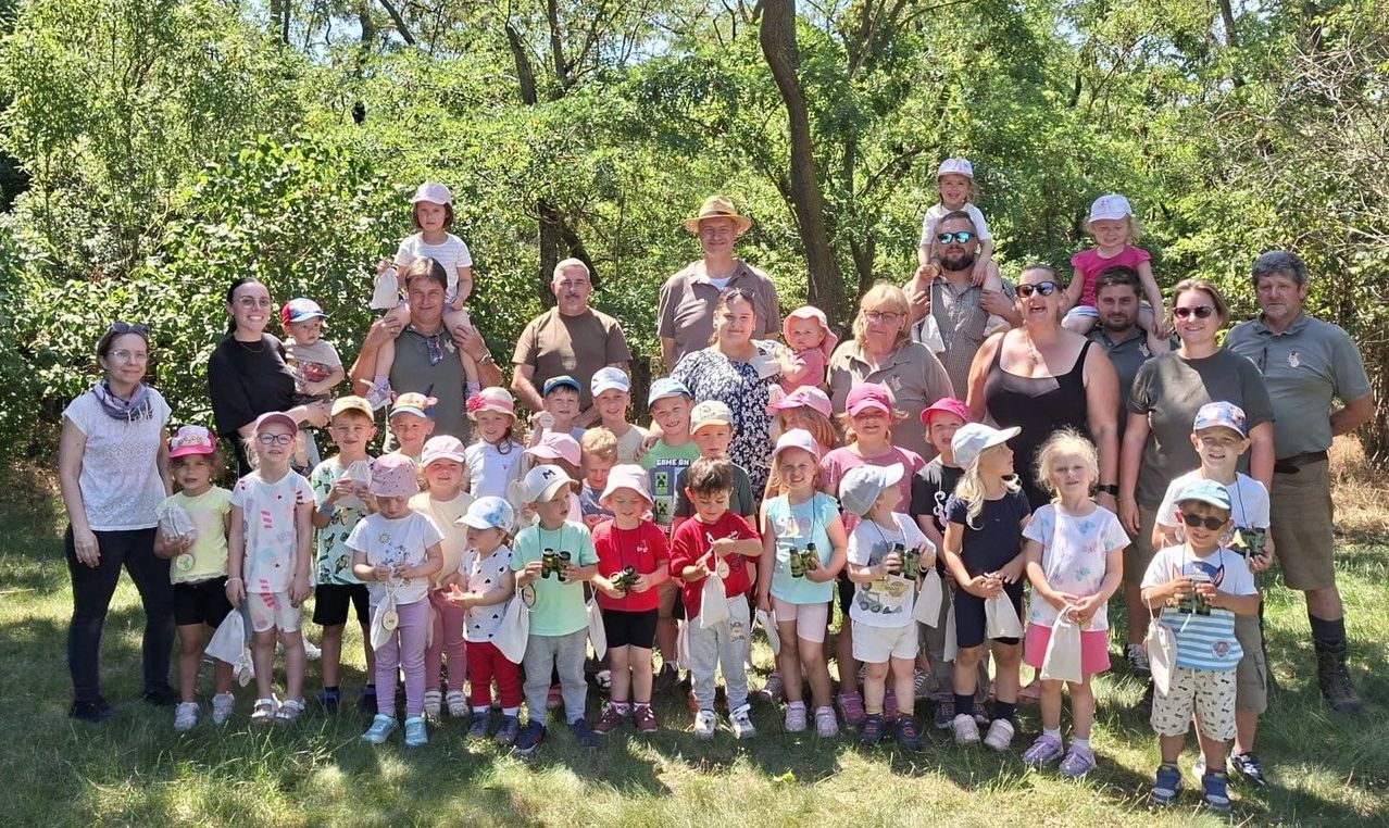 Eine große Gruppe von Kindern und Erwachsenen versammelt sich auf einem Grasfeld. Einige halten Ferngläser in der Hand und die meisten tragen Hüte. Einige Erwachsene stehen hinter den Kindern. Der Hintergrund ist mit Bäumen gefüllt.