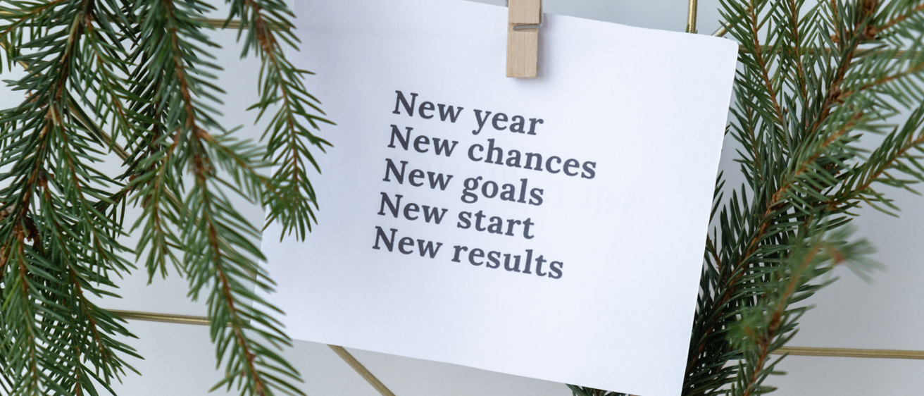 Ein weißes Papier mit einer Liste von motivierenden Phrasen hängt an einem Holzklammer, umgeben von grünen Tannenzweigen. Die Phrasen beinhalten 'Neues Jahr,' 'Neue Chancen,' 'Neue Ziele,' 'Neuer Anfang' und 'Neue Ergebnisse.' Der Hintergrund deutet auf eine festliche, vielleicht weihnachtliche Atmosphäre hin.