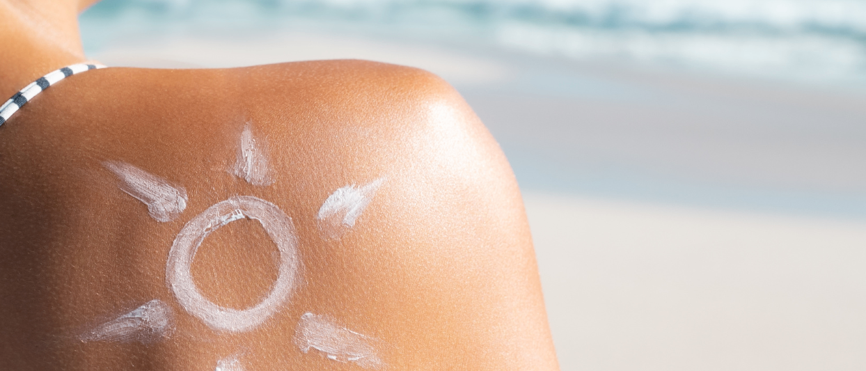 Eine Person steht am Strand mit Sonnencreme auf dem Rücken, dem Ozean zugewandt.