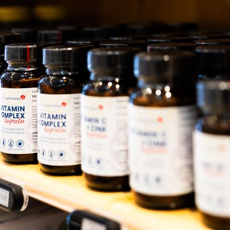 Mehrere Flaschen mit Vitamin Complex Kapseln sind auf einem Regal angeordnet. Jede Flasche hat einen schwarzen Deckel und ein weißes Etikett mit blauem Text. Der Hintergrund ist unscharf.