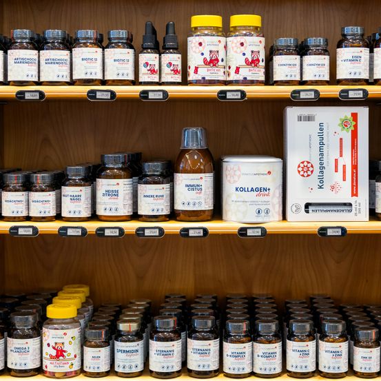 Ein hölzerne Regal zeigt verschiedene Gesundheitsergänzungsmittel, einschließlich Flaschen mit den Aufschriften 'Immune Life', 'Biotic 13' und 'Vitamin C'. Es gibt auch Schachteln mit der Aufschrift 'Kollageneinpumpeln'. Die Flaschen sind ordentlich in Reihen angeordnet, mit sichtbaren Preisschildern auf jeder.