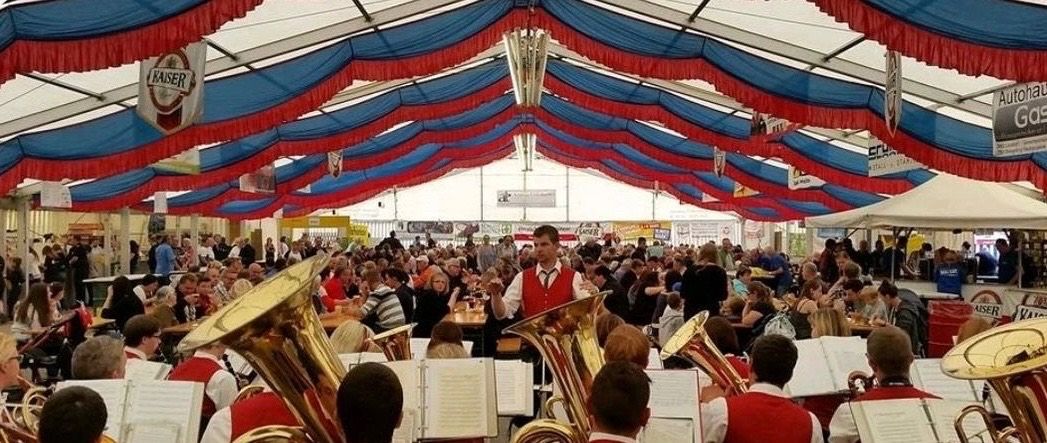 Eine Band spielt Blechblasinstrumente in einem großen Zelt mit blauen und roten Dekorationen. Viele Menschen sind versammelt, einige sitzen und andere stehen.