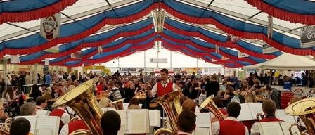Eine Band spielt Blechblasinstrumente in einem großen Zelt mit blauen und roten Dekorationen. Viele Menschen sind versammelt, einige sitzen und andere stehen.