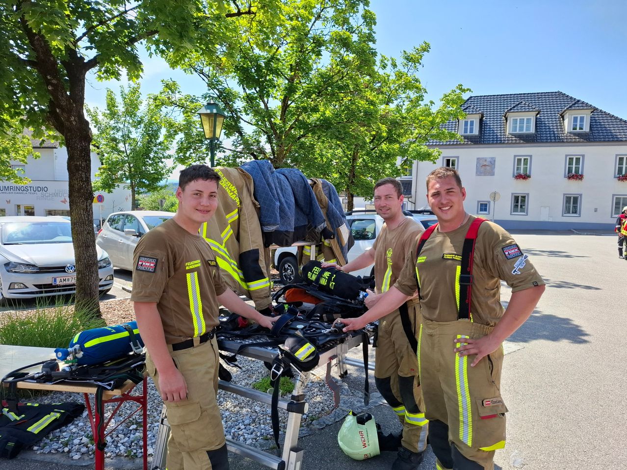 Drei Feuerwehrleute stehen draußen neben Ausrüstung. Sie tragen Uniformen mit reflektierenden Streifen. Hinter ihnen ist ein Gebäude mit grünem Dach zu sehen. Bäume und geparkte Autos sind ebenfalls im Bild.