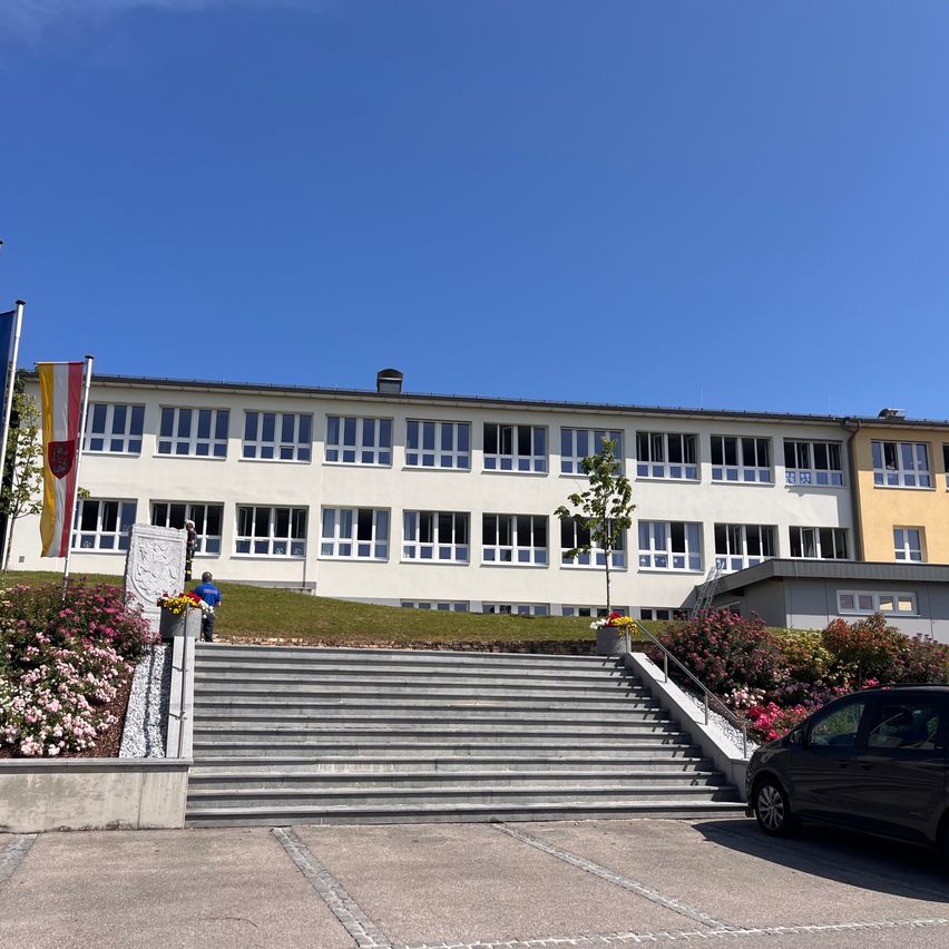 Ein großes weißes Gebäude mit vielen Fenstern und Treppen, die hinaufführen. Eine Person in einem blauen Shirt ist auf dem Rasen. Ein Auto ist auf dem Parkplatz geparkt.