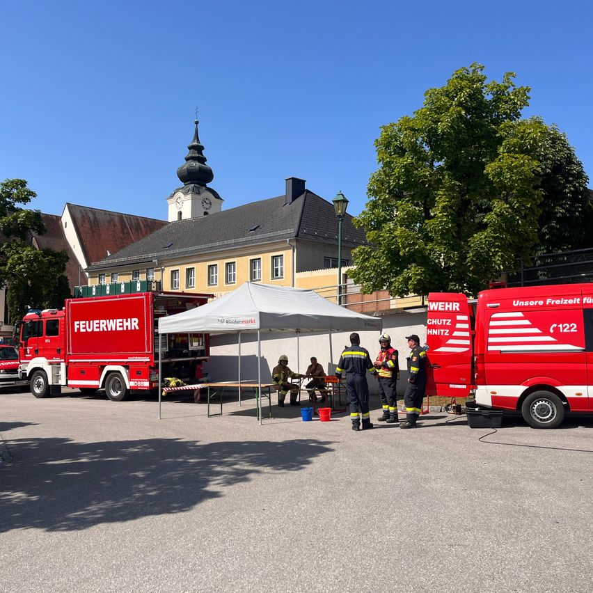 Feuerwehrleute stehen um einen Tisch unter einem Zelt vor einem Feuerwehrauto. Ein rotes Fahrzeug mit der Nummer 122 ist daneben geparkt. Im Hintergrund befindet sich ein Turm mit einer Uhr.