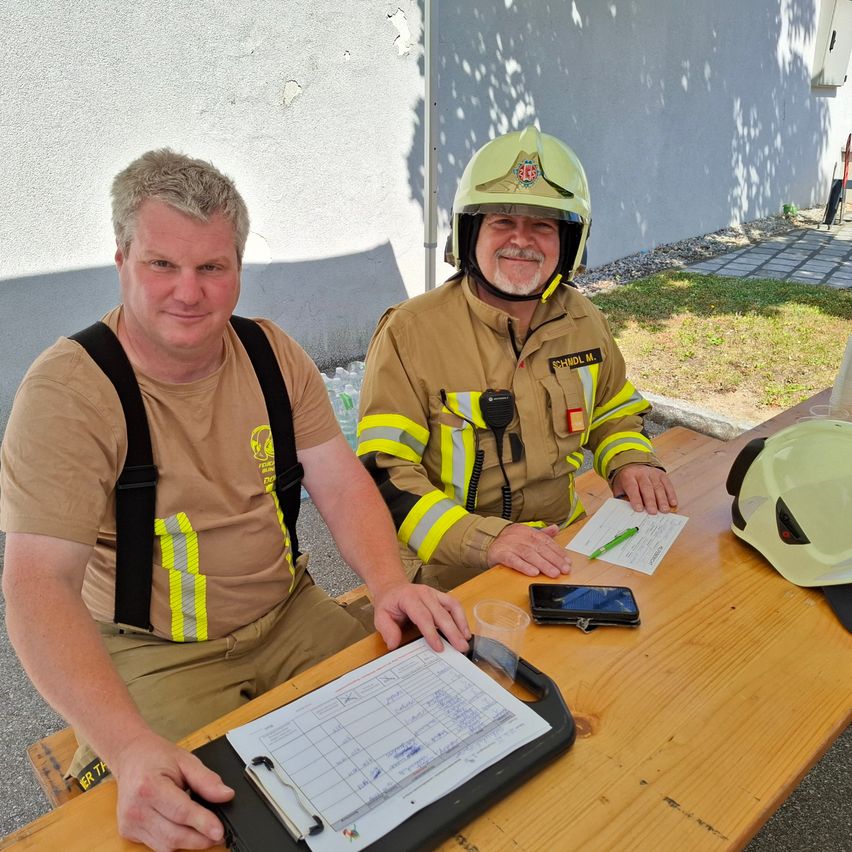 Zwei Feuerwehrleute in Uniform sitzen lächelnd für die Kamera an einem Tisch im Freien. Einer hält ein Papier, während der andere einen Helm trägt. Ein Telefon, ein Stift und andere Gegenstände liegen auf dem Tisch.