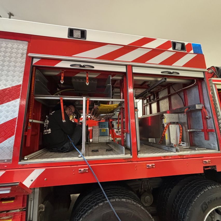 Ein Feuerwehrmann arbeitet in einem roten Feuerwehrwagen an der Ausrüstung. Die Türen des Wagens sind geöffnet, und verschiedene Werkzeuge und Geräte sind im Inneren sichtbar.