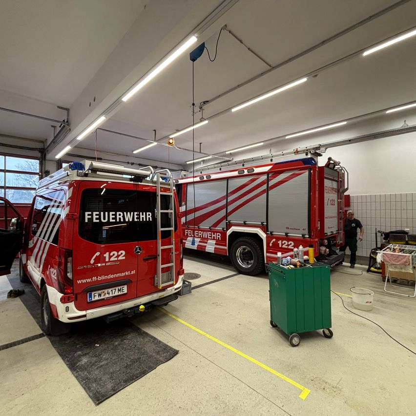 Zwei rote Feuerwehrfahrzeuge sind in einer Garage geparkt. Eines hat die Nummer 122 auf der Seite. Ein Mann steht neben dem Fahrzeug, und ein grüner Trolley ist in der Nähe.