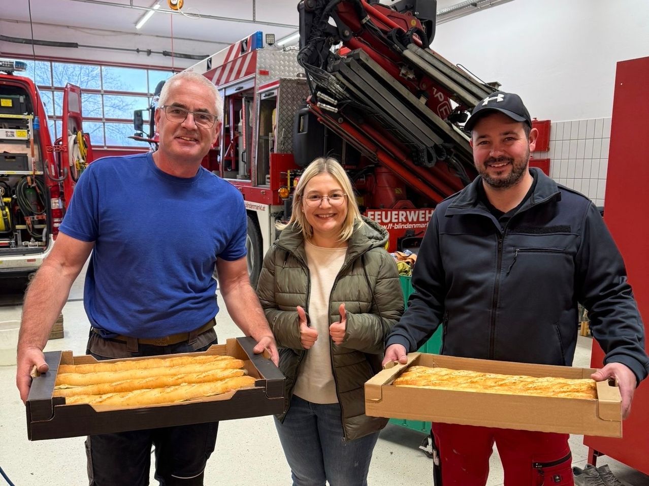 Drei Menschen stehen in einem Raum, lächeln und posieren für ein Foto. Zwei Männer halten Brotboxen und eine Frau in der Mitte gibt einen Daumen hoch. Ein Feuerwehrwagen ist hinter ihnen geparkt.