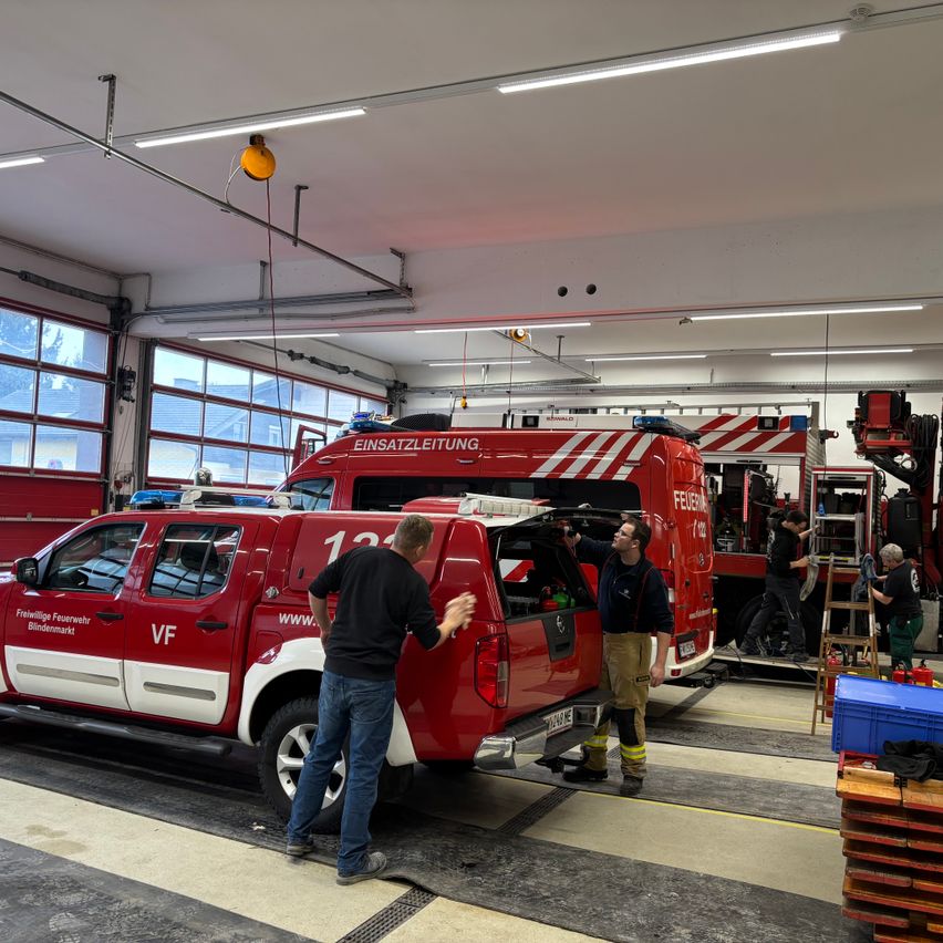 In einer Feuerwehrwache inspizieren zwei Feuerwehrleute einen roten Pickup-Truck mit der Nummer 12 an der Seite. Andere Mitarbeiter sind mit verschiedenen Aufgaben beschäftigt, und Feuerwehrfahrzeuge sind im Hintergrund geparkt.
