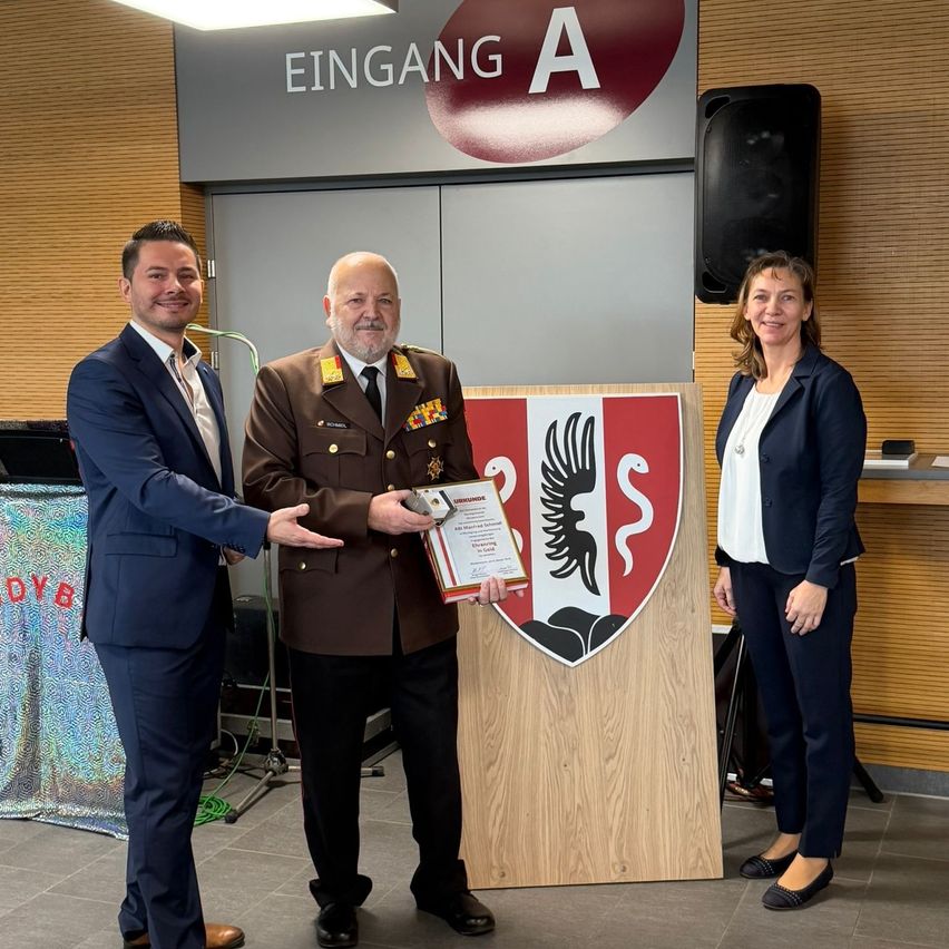 Drei Personen stehen vor einem Holzschild, ein Mann in Uniform hält ein Zertifikat, ein anderer Mann schüttelt seine Hand und eine Frau steht neben ihnen.